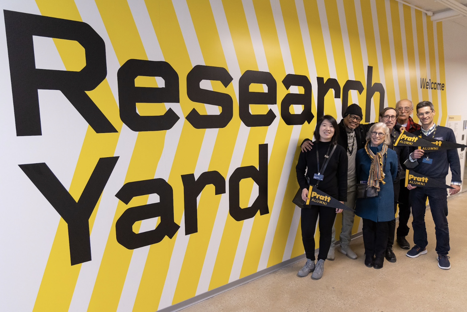 A group of six people standing together in front of a large yellow and white striped wall with the words "Research Yard" in bold black letters. They are holding black and yellow "Pratt Alumni" pennants and smiling at the camera. The group is casually dressed, and the wall behind them gives the space a modern and vibrant feel.