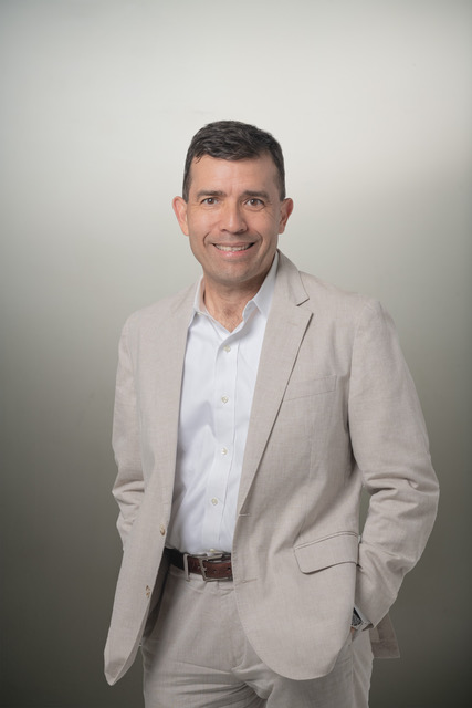 A man with short dark hair, wearing a light gray suit jacket over a white dress shirt. He is smiling at the camera while posing with his hands in his pockets, standing against a plain light gray background.