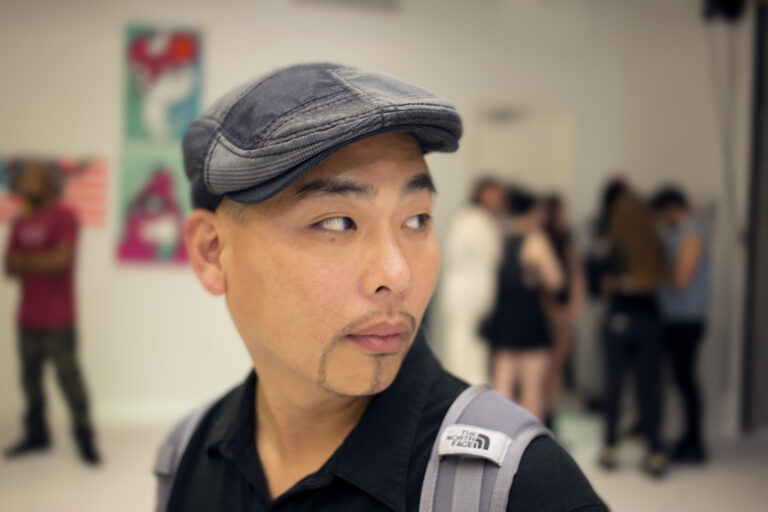 A man with a light goatee and short hair, wearing a gray flat cap and a black shirt with a North Face backpack. He is looking off to the side in a crowded gallery or art space, with blurred people in the background.