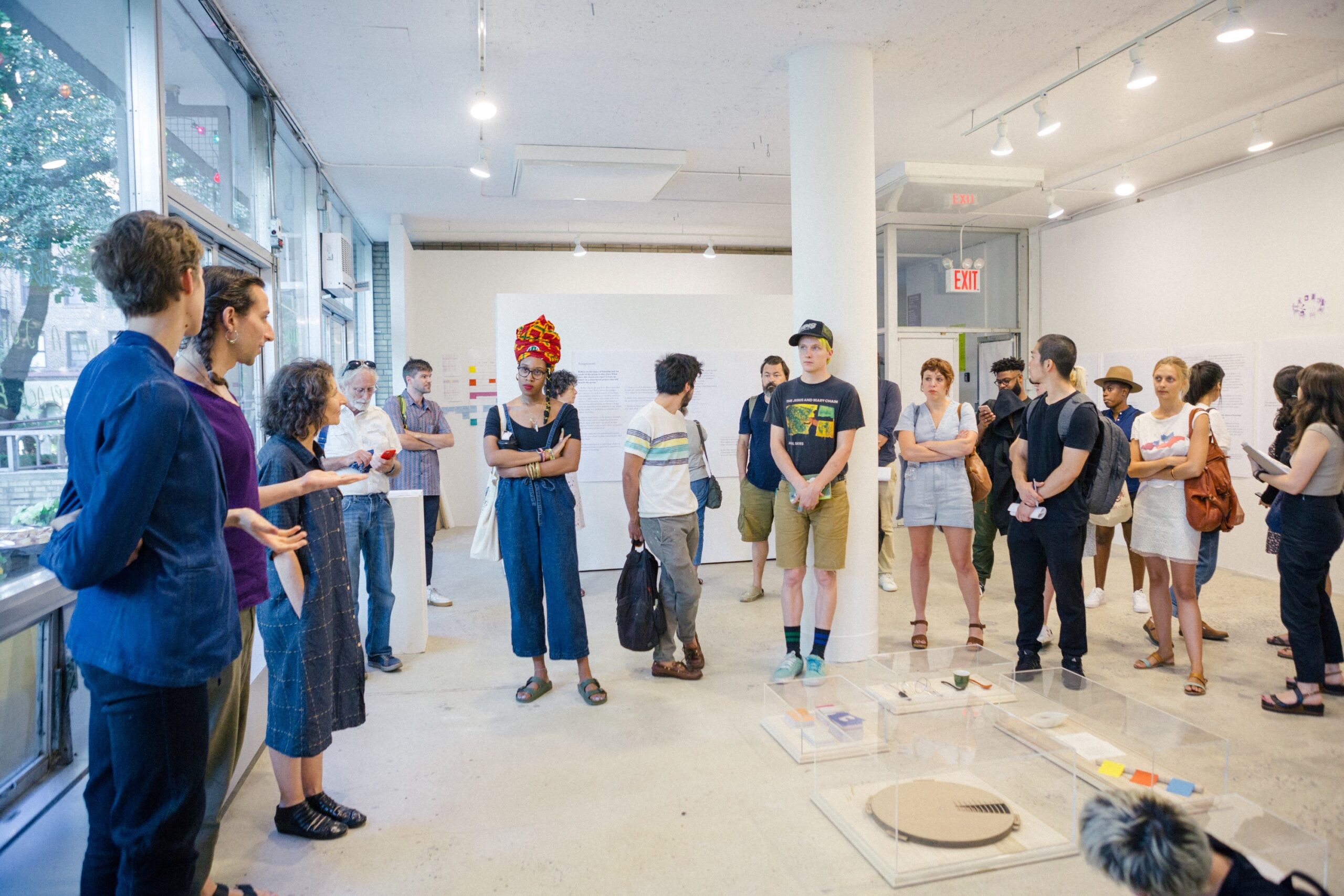 Groups of people in a room with concrete floor and white walls, resembling a gallery, load bearing post in the middle of room