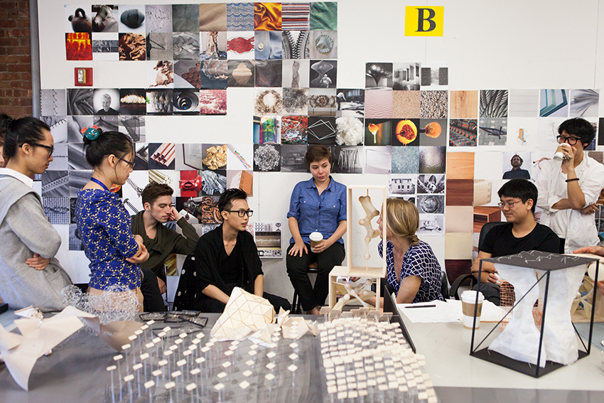 A group of students and a professor engage in a discussion around a table covered with architectural models and design prototypes. The background wall is filled with a collage of various images and materials.