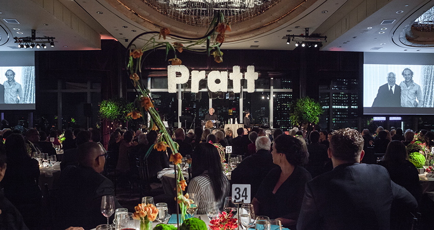 A large gathering at a formal event, with attendees seated at round tables adorned with flowers and candles. At the front, a speaker is addressing the audience from a podium, with a large "Pratt" sign in the background. Two large screens display a black and white photo of an older couple. The venue is elegantly decorated, with chandeliers and cityscape views visible through the windows.