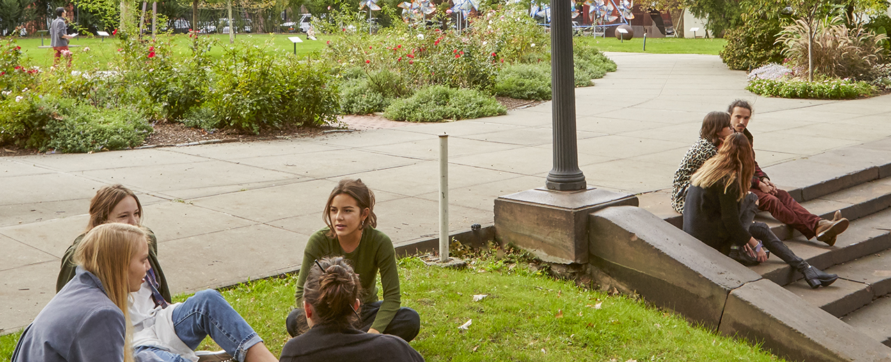 Students sit on grass and on stairs in a park as they chat casually.