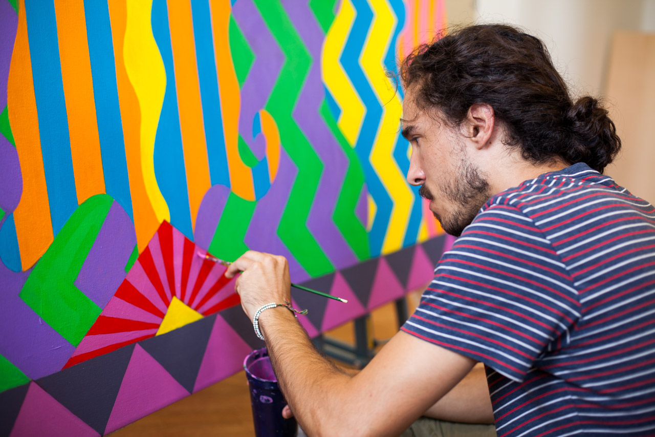 A student crouches in front of a canvas with brightly-colored geometric vertical shapes. He is painting on the canvas.