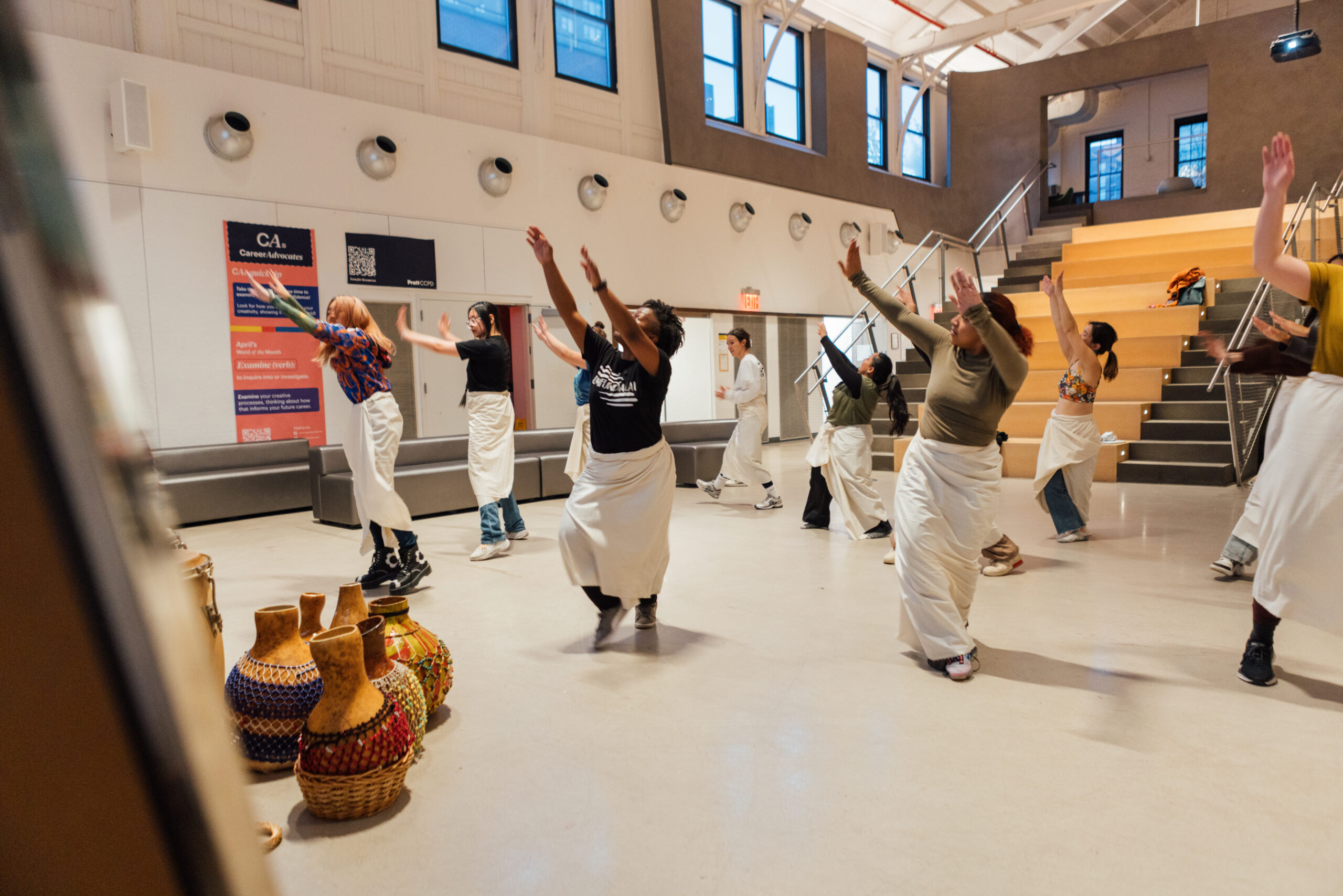 A group of people participates in a traditional dance class in a brightly lit indoor space with high ceilings and modern architecture. The dancers wear white wrap skirts over their clothing and are mid-movement, arms raised, following choreography. In the foreground, several shekeres—African percussion instruments—rest on the floor, suggesting a cultural or rhythmic component to the session. The environment appears to be a university or community center with amphitheater-style seating in the background.